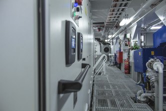 Interior corridor of a ship’s engine or machinery room with a narrow walkway made of metal grating running down the center.