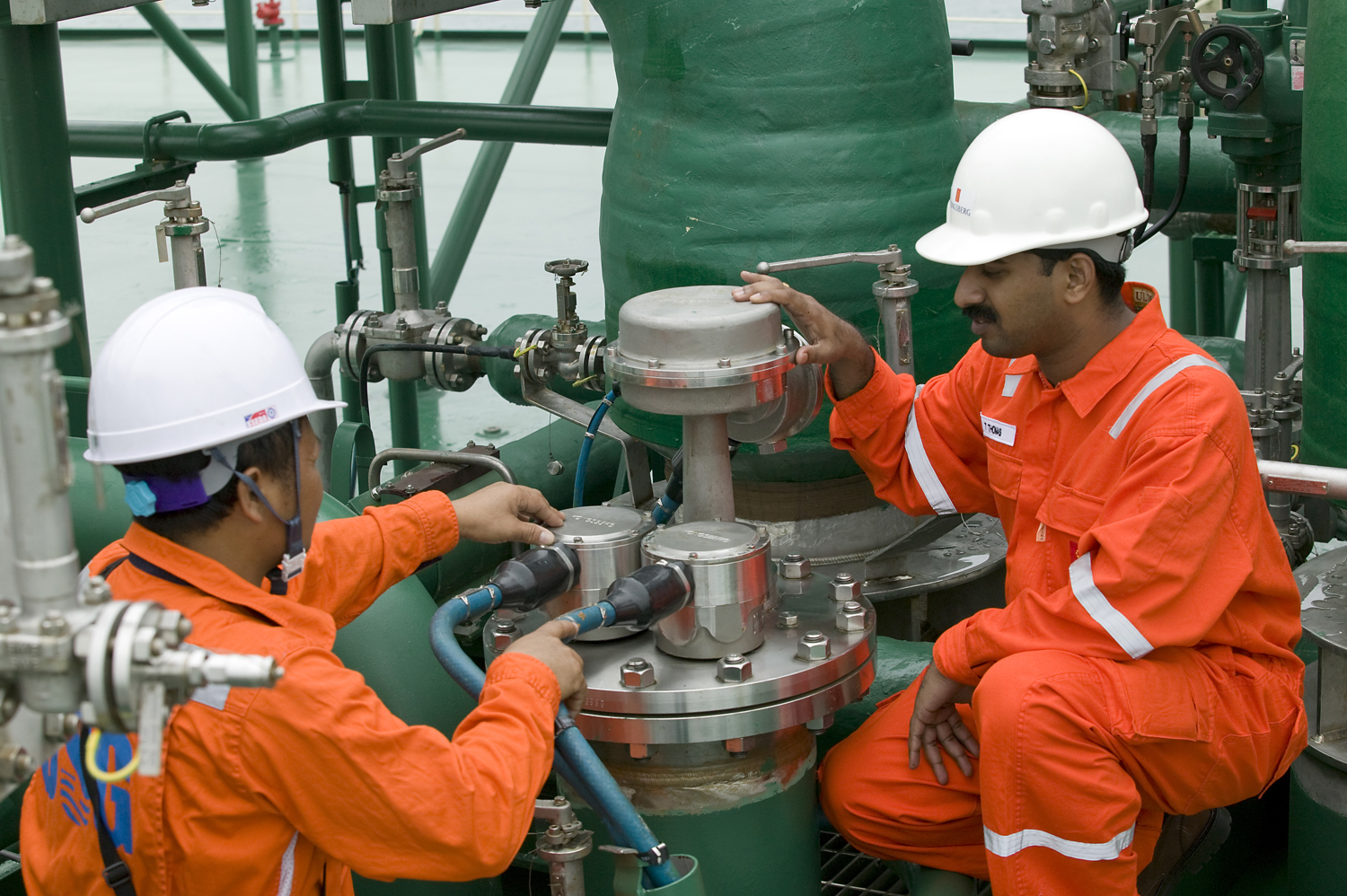 Two offshore workers wearing bright orange protective coveralls and white hard hats are performing maintenance on a large green industrial pipe system