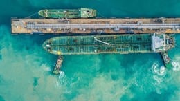 Top-down aerial view of a large tanker moored alongside a long industrial dock, both stretching horizontally across turquoise-green water.