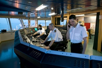 Interior of a ship’s bridge with three crew members operating navigation and control consoles.