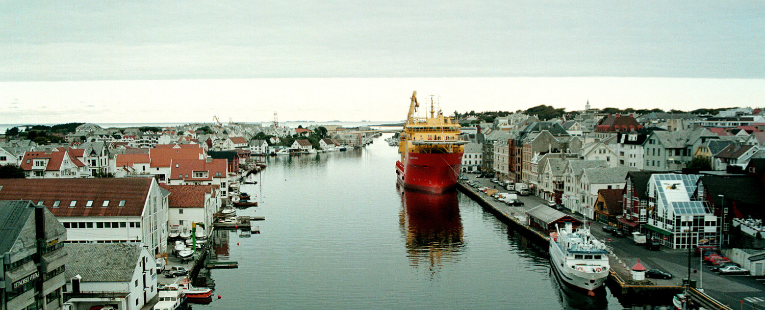 Aerial view of a red offshore vessel navigating a narrow canal through a coastal town.