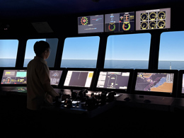 Interior of a modern ship’s bridge showing a person standing at a central control console, facing forward toward large panoramic windows overlooking calm open sea and a clear horizon